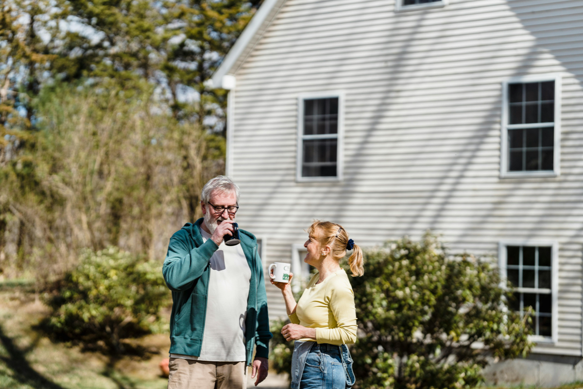 Older man and woman standing outside a white house, smiling and drinking from mugs on a sunny day.